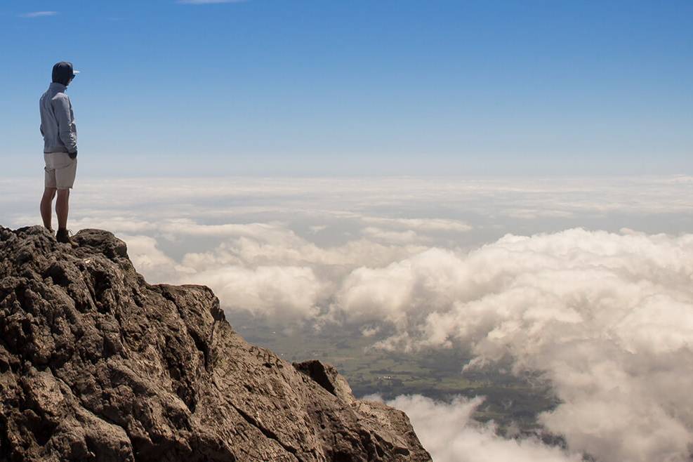 Sinnbild für eine klare, eindeutige Positionierung eines Unternehmens am Markt und für eine deutliche Abgrenzung vom Wettbewerb. Das Bild zeigt einen einzelnen Menschen auf dem Gipfel eines Berges, unter ihm eine Wolkendecke.