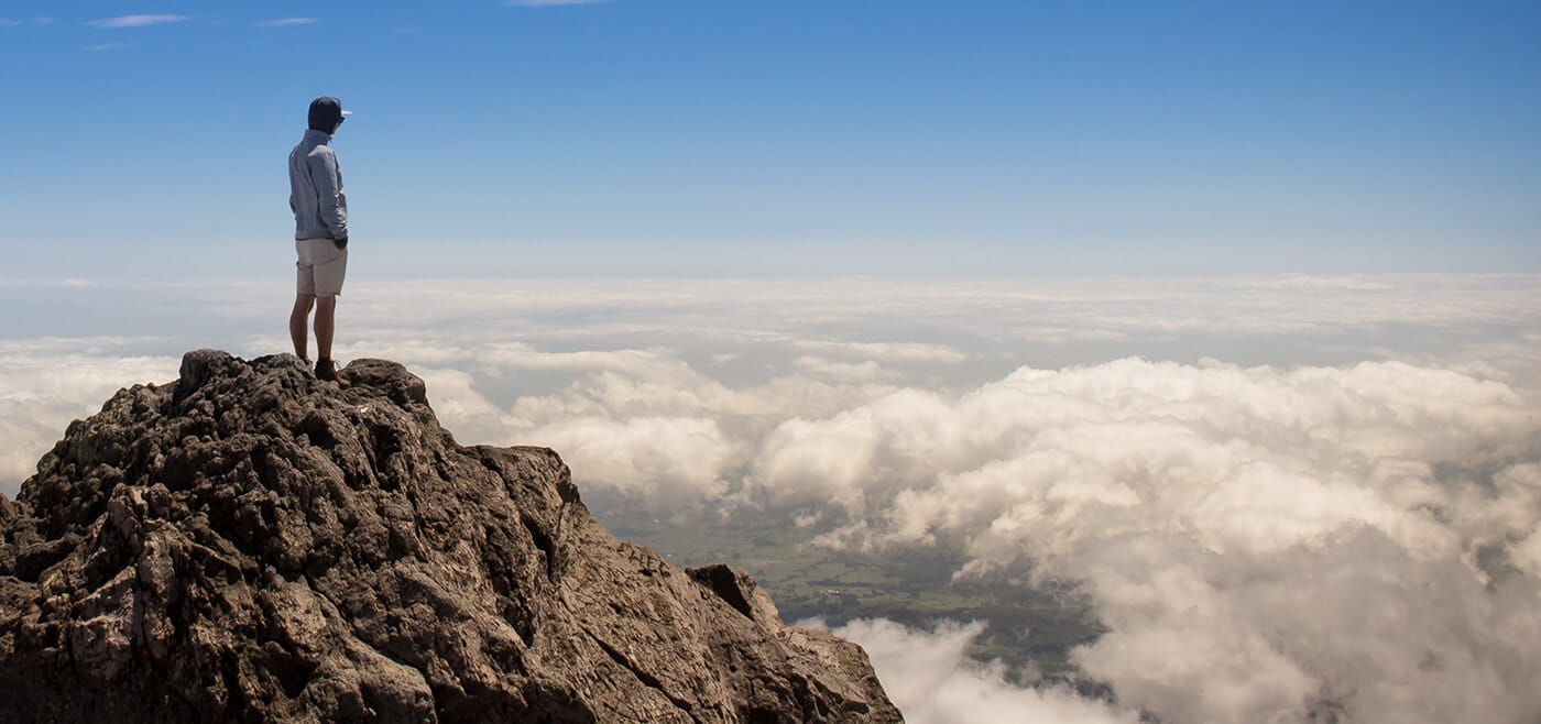 Sinnbild für eine klare, eindeutige Positionierung eines Unternehmens am Markt und für eine deutliche Abgrenzung vom Wettbewerb. Das Bild zeigt einen einzelnen Menschen auf dem Gipfel eines Berges, unter ihm eine Wolkendecke.